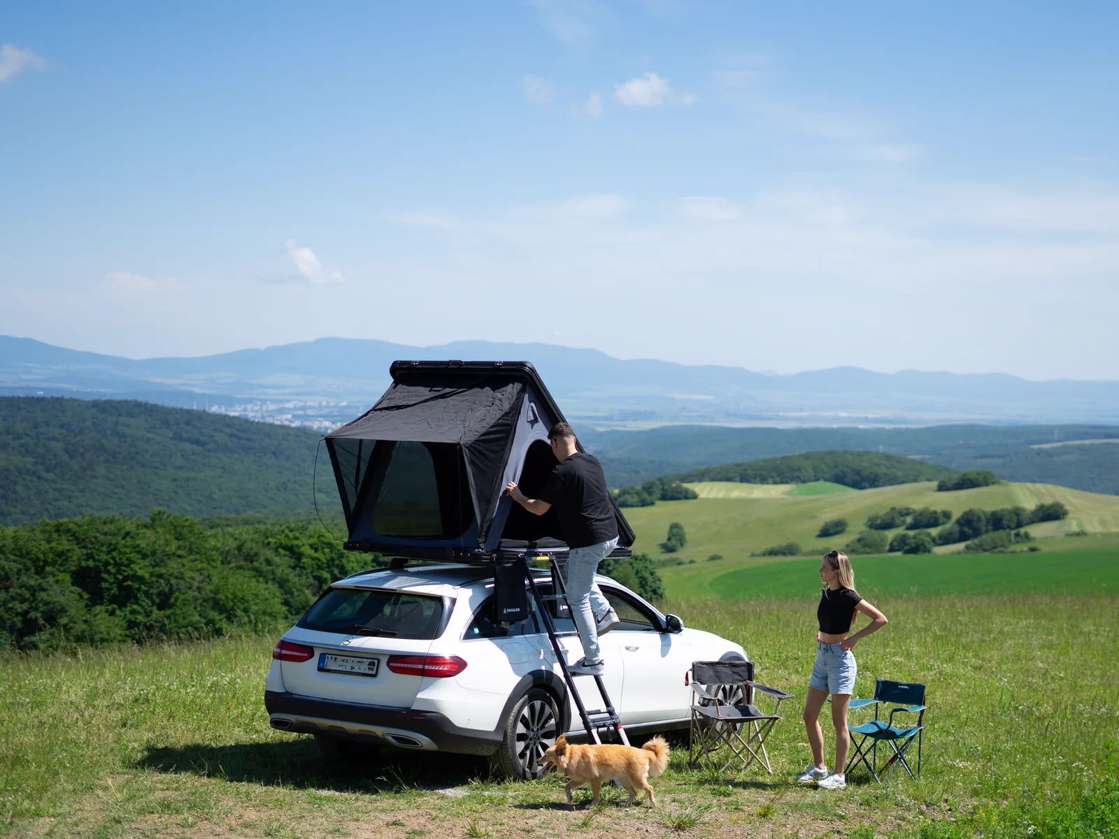 Couple setting up a SNAILER roof tent on a car in a mountain meadow